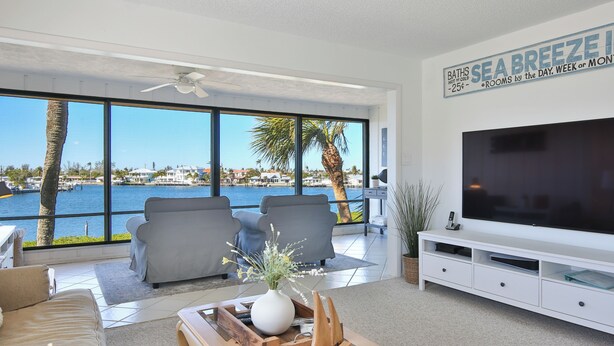 A modern living room with a large TV, grey sofas, a wooden coffee table, and a view of the sea. - Sam's Cove! Second Floor Condo Overlooking Water (Holmes Beach)