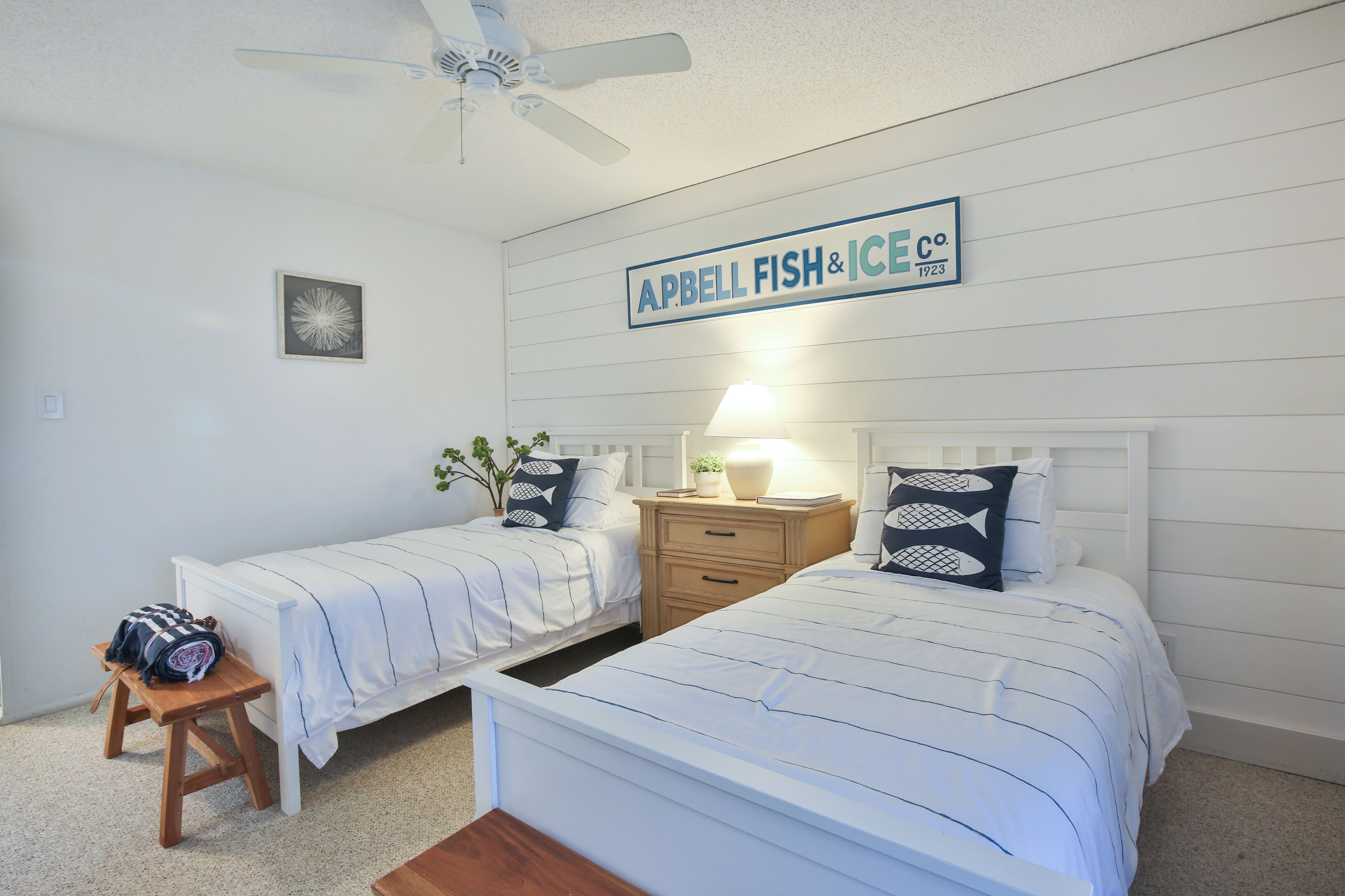A bedroom with two beds, a ceiling fan, a sign above the beds, and a small table with a bag.
