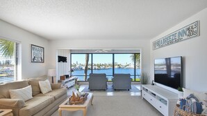 A modern living room with a large TV, comfortable seating, a view of the sea, and a sign advertising meals at all hours. - Sam's Cove! Second Floor Condo Overlooking Water (Holmes Beach)