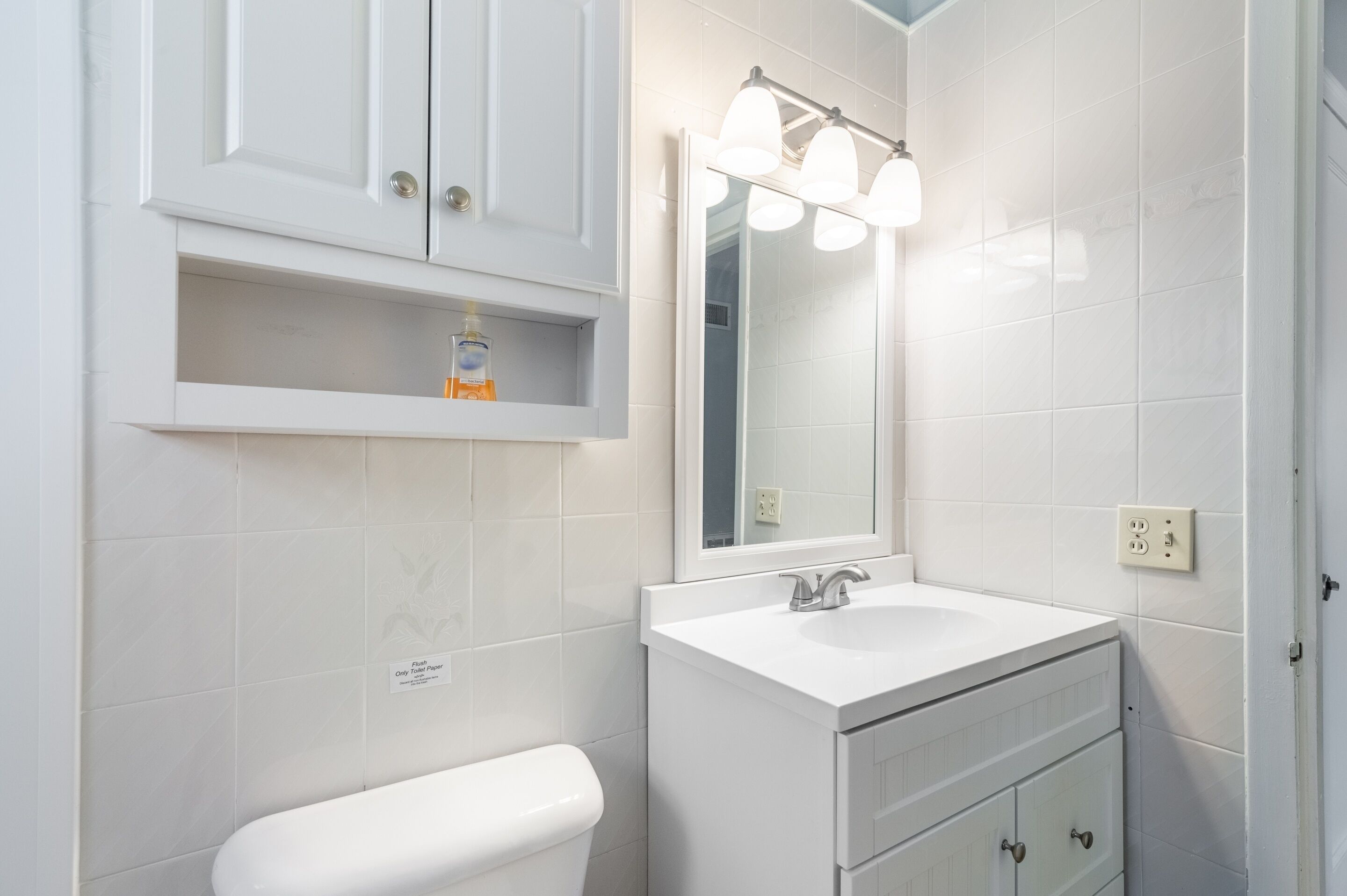 A bathroom with a white toilet, a sink with a mirror above it, and a shelf above the sink.