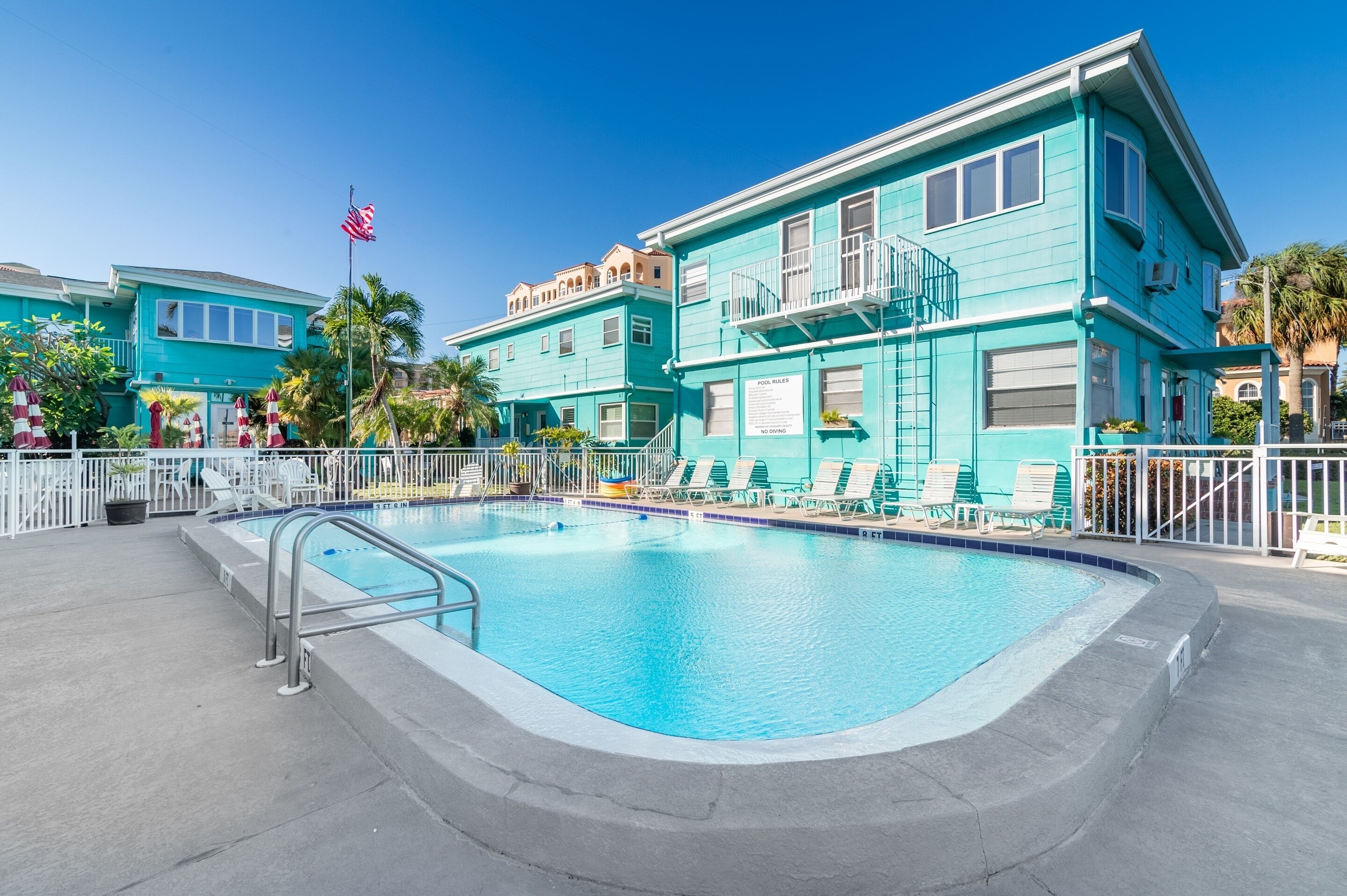 A turquoise building with a pool, lounge chairs, and a flag.