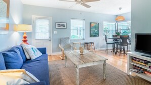 Ein Wohnzimmer mit einem blauen Sofa, einem Holztisch, einem Deckenventilator und einem Fernseher auf einem weißen Schrank.