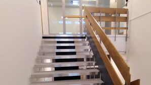 A staircase with metal steps and a wooden handrail, leading to a room with a glass door and a visible emergency exit sign. - Hotelli Kerava (Kerava)