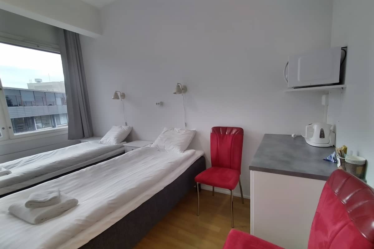 Habitación de hotel con dos camas individuales, una silla roja, un escritorio pequeño con una tetera y una ventana con cortinas.