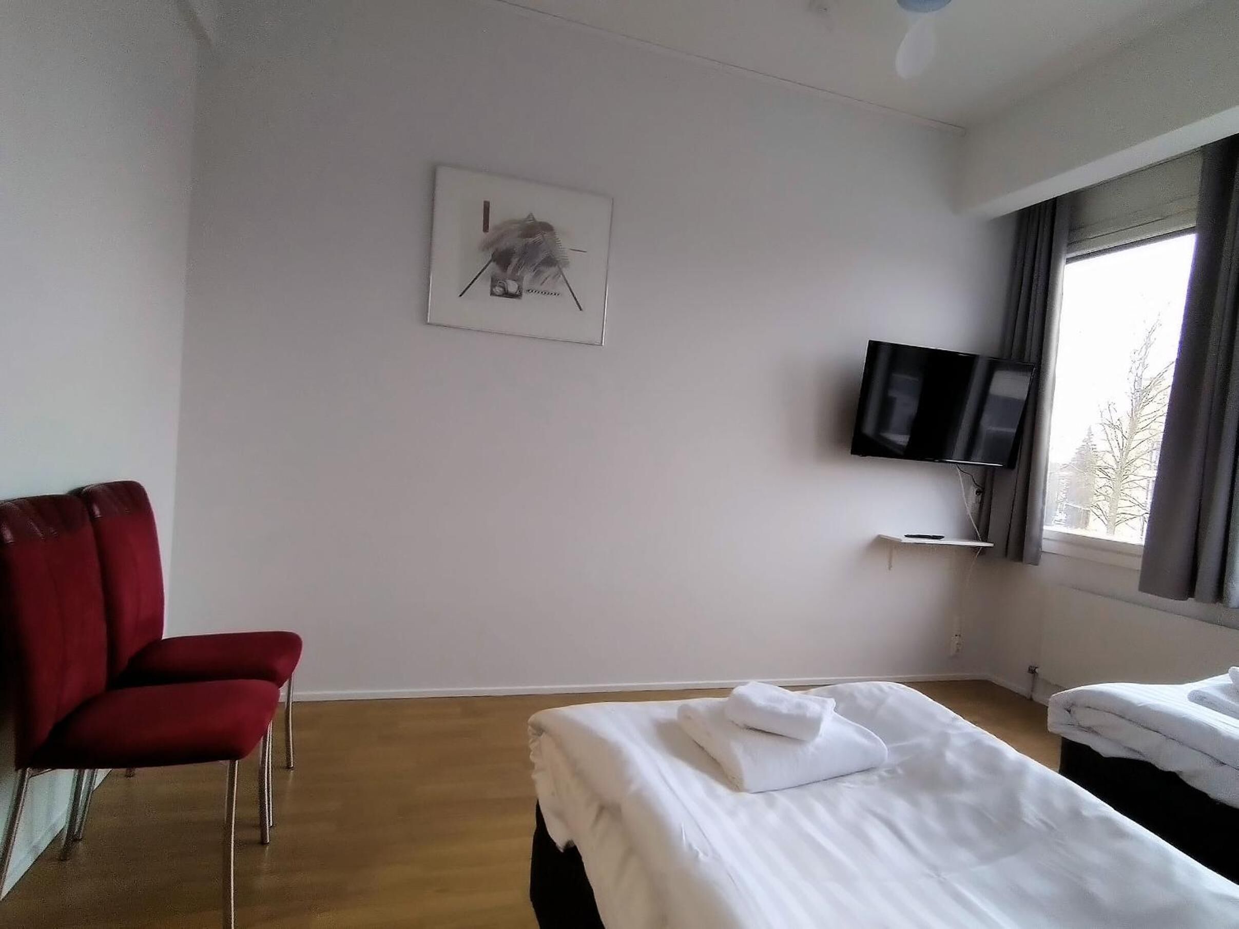 Habitació d'hotel amb un llit, una cadira vermella, una televisió i una finestra que mostra arbres.