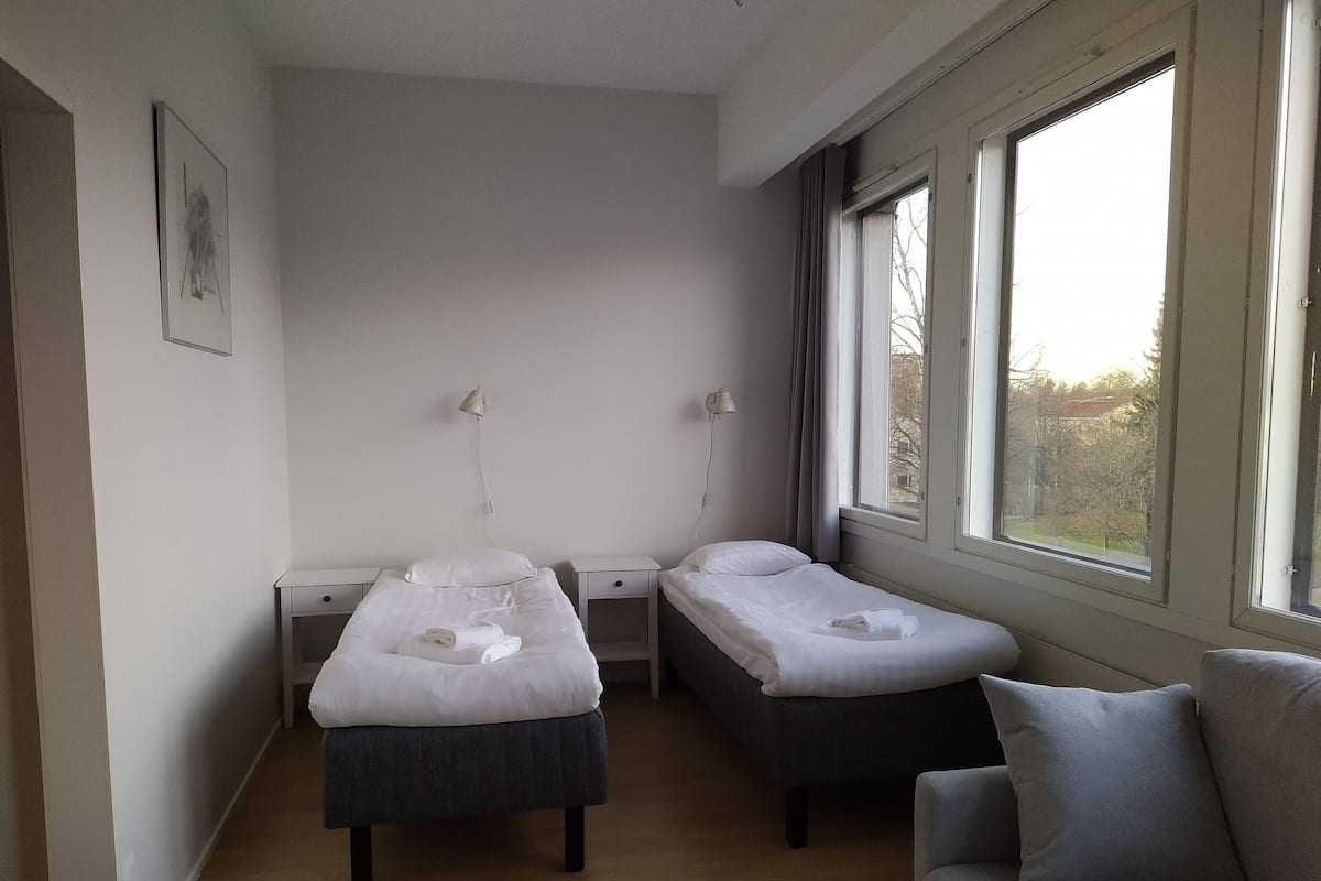 Una habitación pequeña y limpia con dos camas individuales, un escritorio, una silla y ventana que ofrece vistas al exterior.