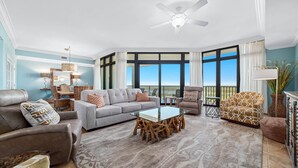 Ein geräumiges Wohnzimmer mit grauem Sofa, braunen Sesseln, einem Glastisch und einem Holztisch. Große Fenster bieten Blick ins Freie.