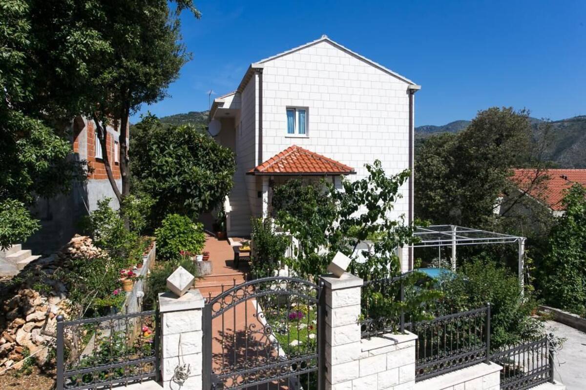 A white two-story house with a red-tiled roof, surrounded by a garden with various plants and a metal gate.