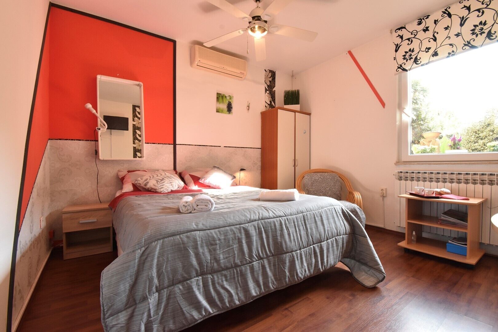 A bedroom with a bed, a nightstand, a chair, a wardrobe, a radiator, and a window with a view of greenery.