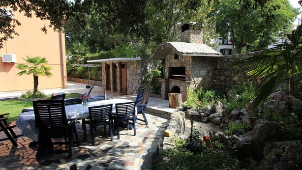Un patio al aire libre con mesas y sillas de piedra, un horno de piedra y abundante vegetación.