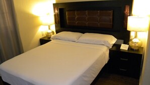 A hotel room with a large bed, two bedside tables with lamps, a dark headboard, and a small sign on one of the tables.
