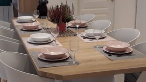Une table à manger dressée avec des assiettes roses, des bols blancs et des verres à vin, entourée de chaises blanches.