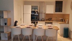 Une cuisine compacte avec des meubles blancs, un four encastré et un coin repas comprenant quatre chaises blanches et des assiettes roses, le tout disposé pour quatre personnes.