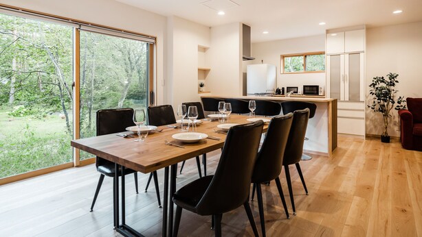 A modern dining area with a wooden table and black chairs, a kitchen in the background, and a large sliding glass door leading to a garden. - Villars Chalet (Hakuba)