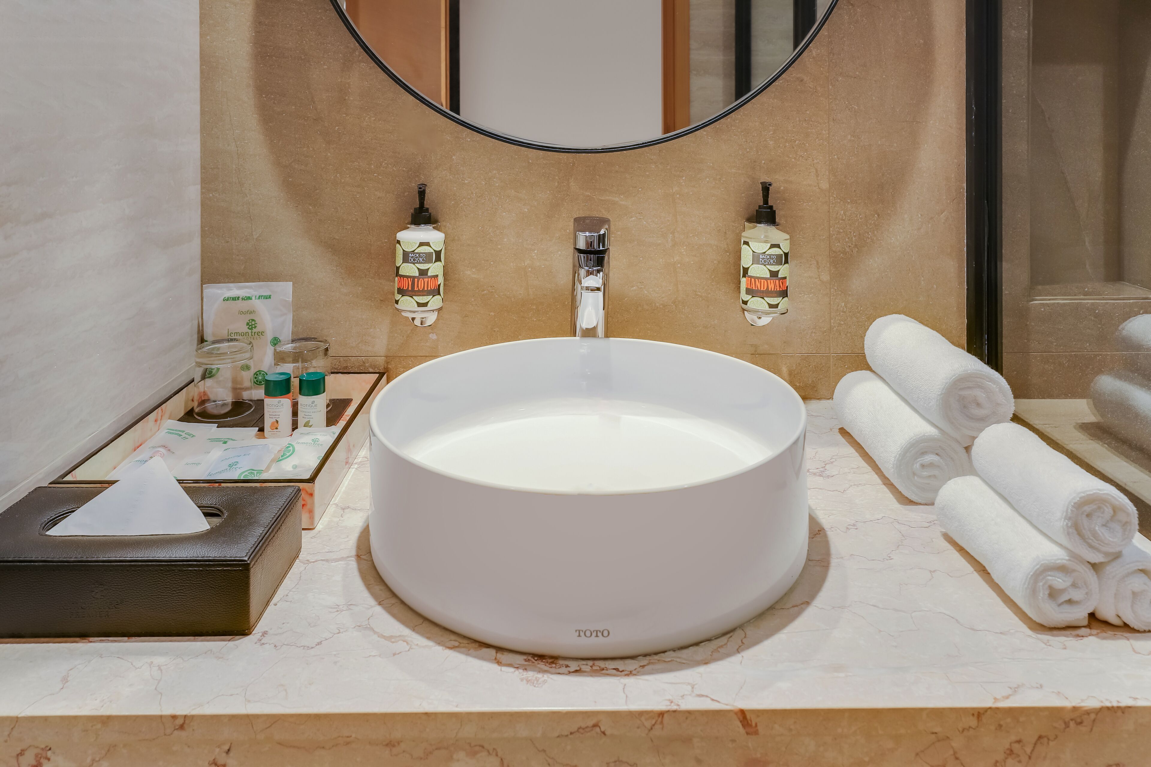 A bathroom with a round white sink, two bottles of hand sanitizer, a tissue box, and rolled towels.