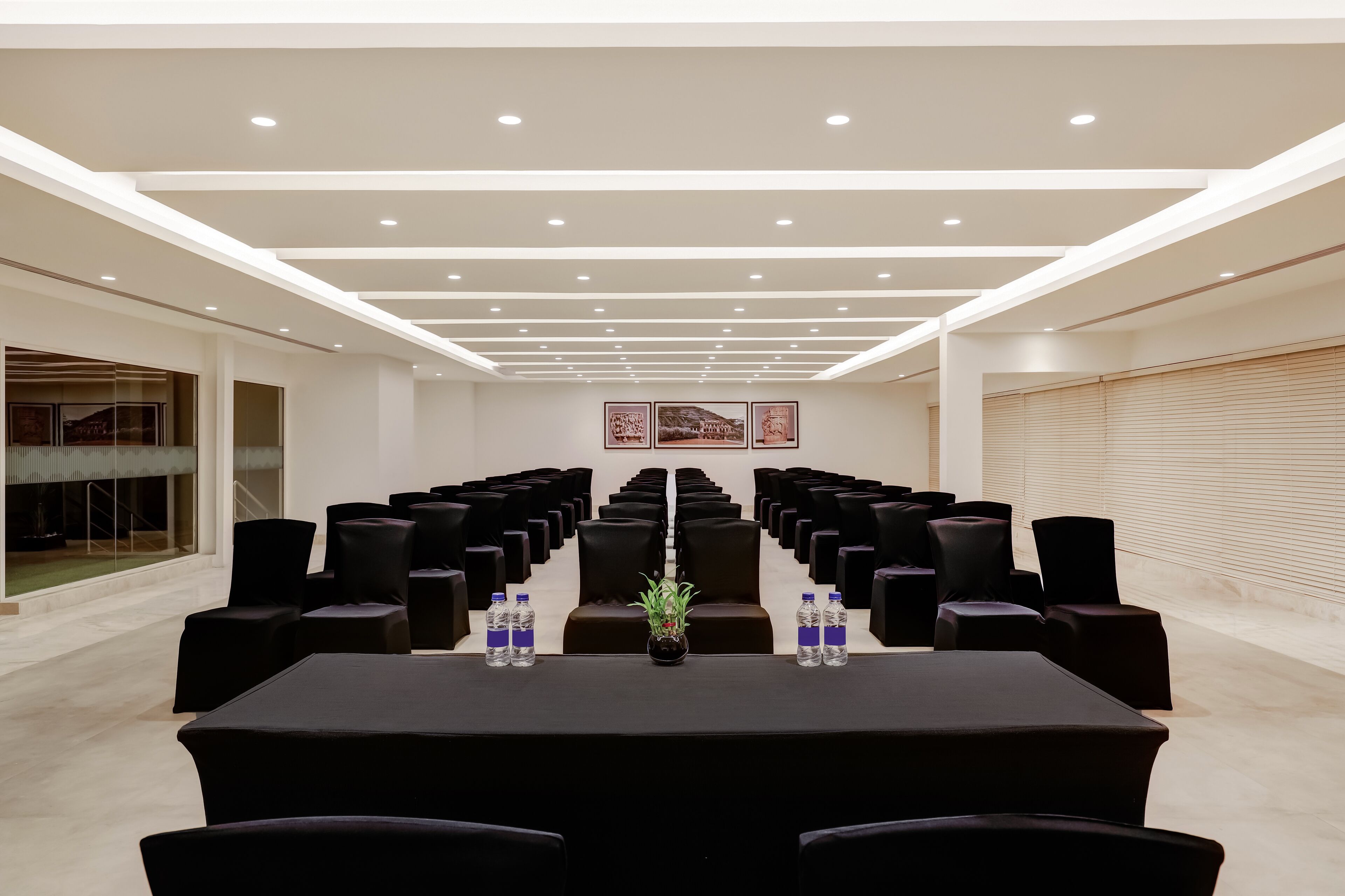 Uma sala de conferência com fileiras de cadeiras pretas, uma mesa comprida com garrafas de água e um vaso de planta.