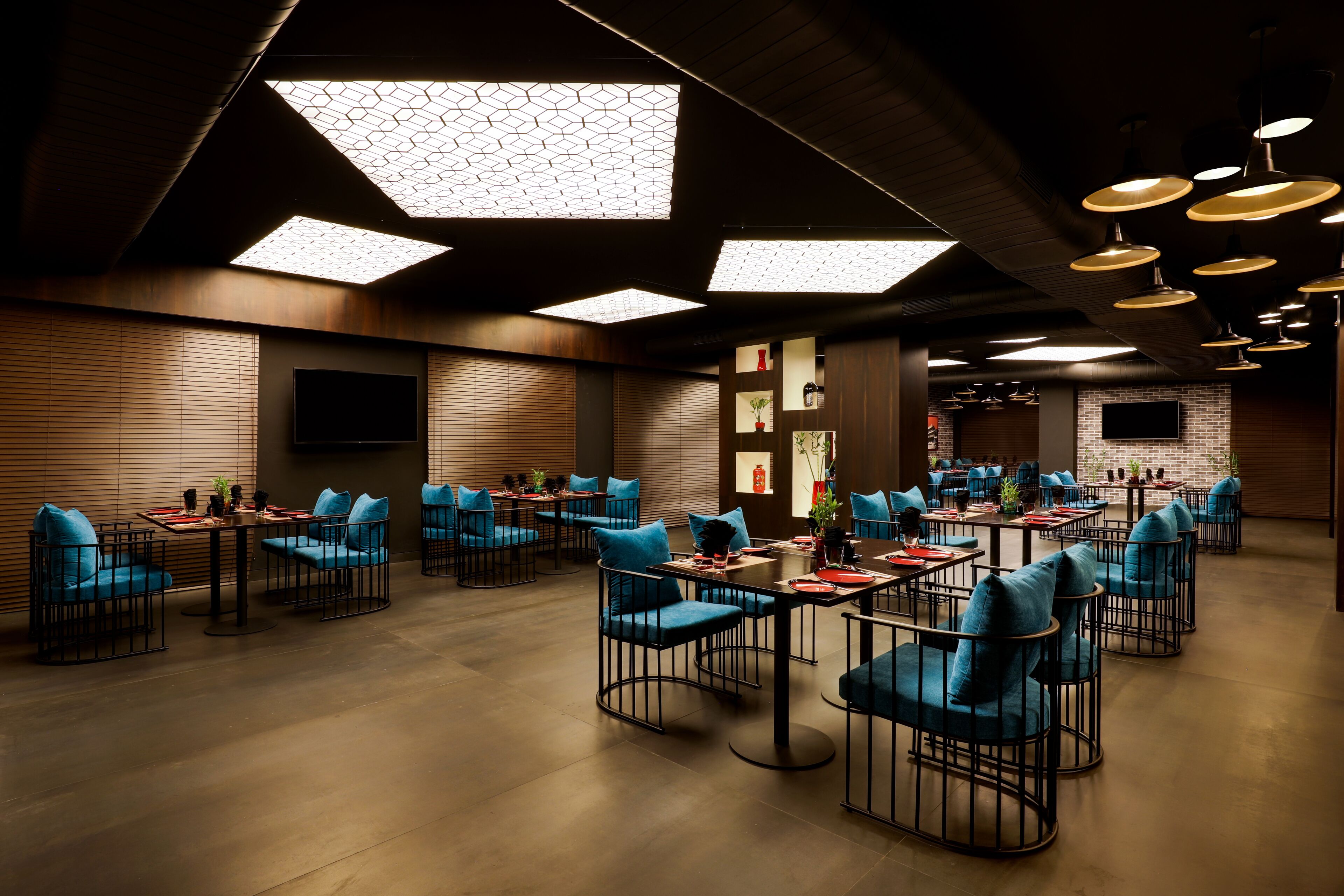 Un restaurant moderne avec un plafond haut, des luminaires au design géométrique et un mur en briques. Les tables sont décorées de chaises bleues et de sets de table rouges.