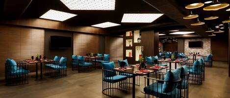 Un restaurant moderne avec un plafond haut, des luminaires au design géométrique et un mur en briques. Les tables sont décorées de chaises bleues et de sets de table rouges.