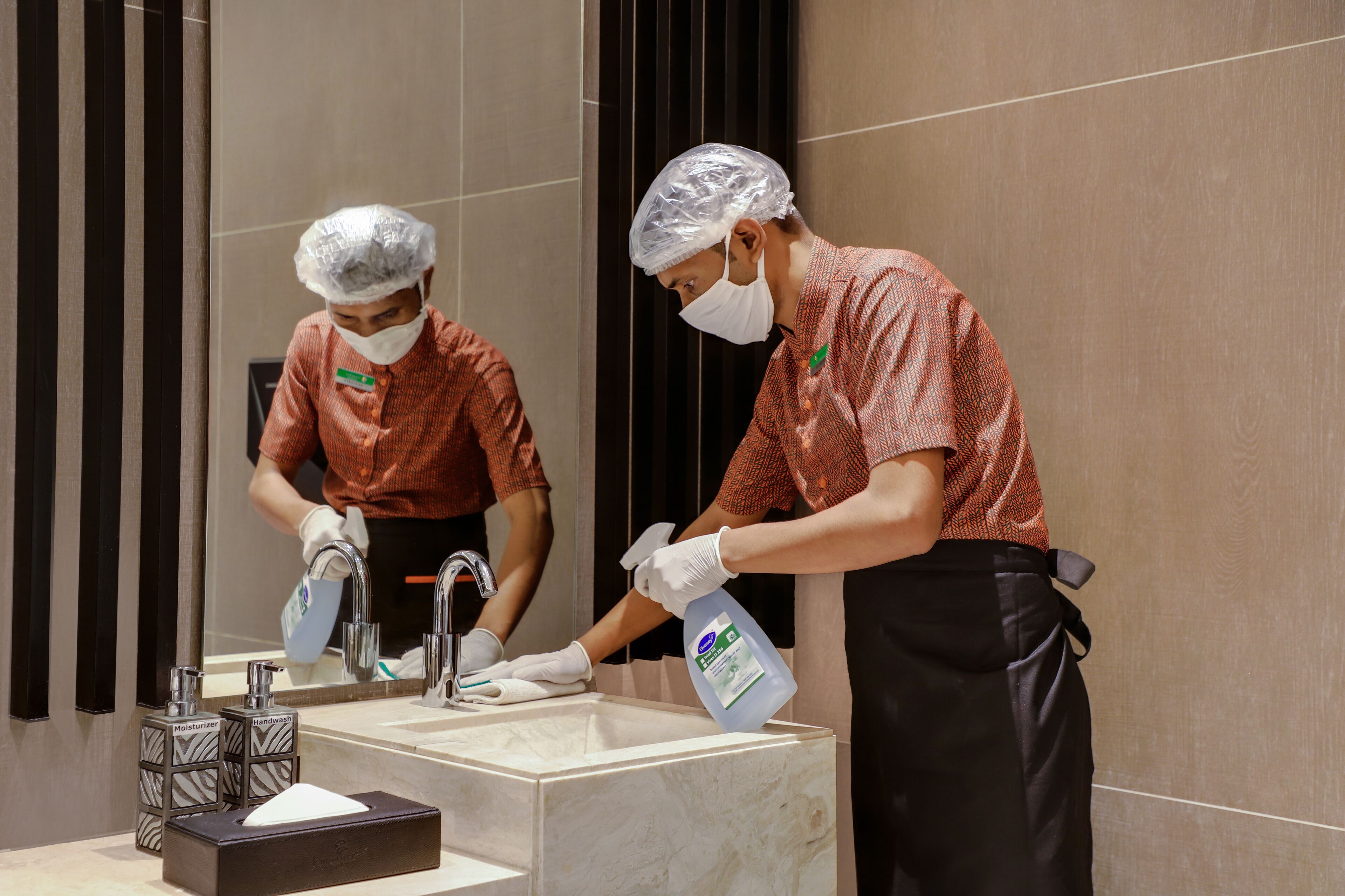 Two individuals cleaning a bathroom sink with a spray bottle and a tissue box.
