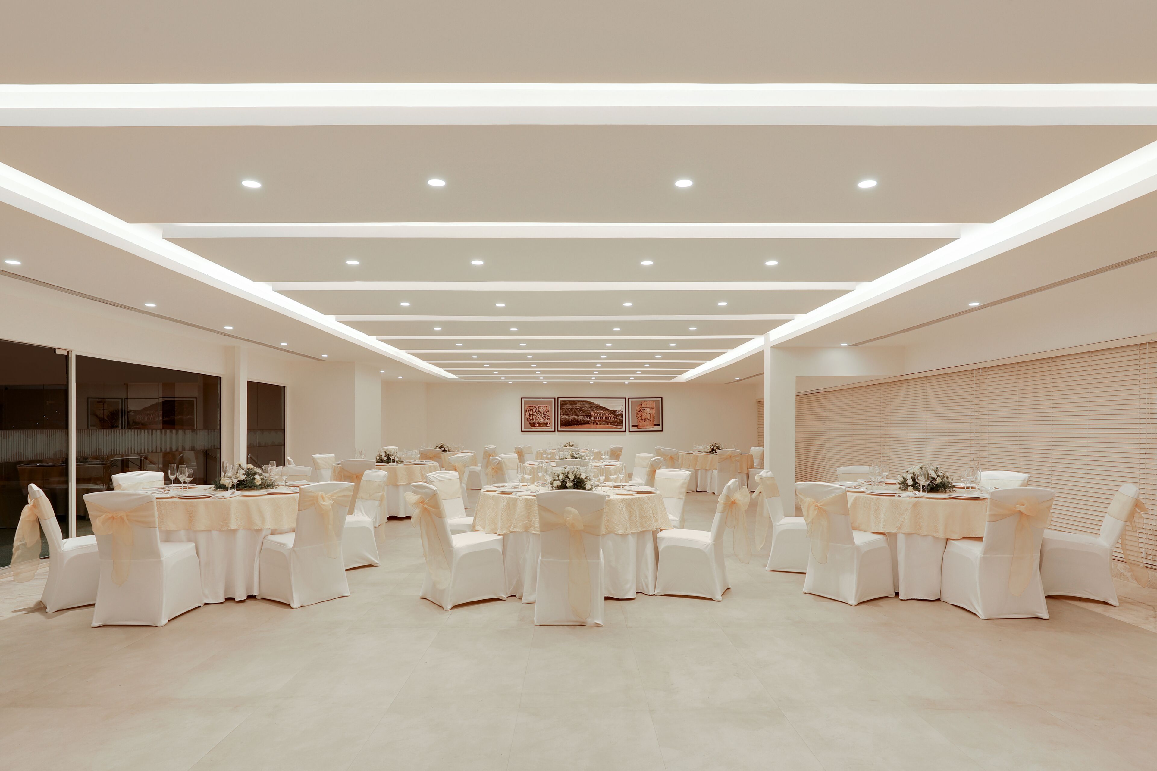 Een ruime balzaal met meerdere ronde tafels, elk gedekt met witte tafelkleden en stoelen, en een modern plafondontwerp met inbouwverlichting.