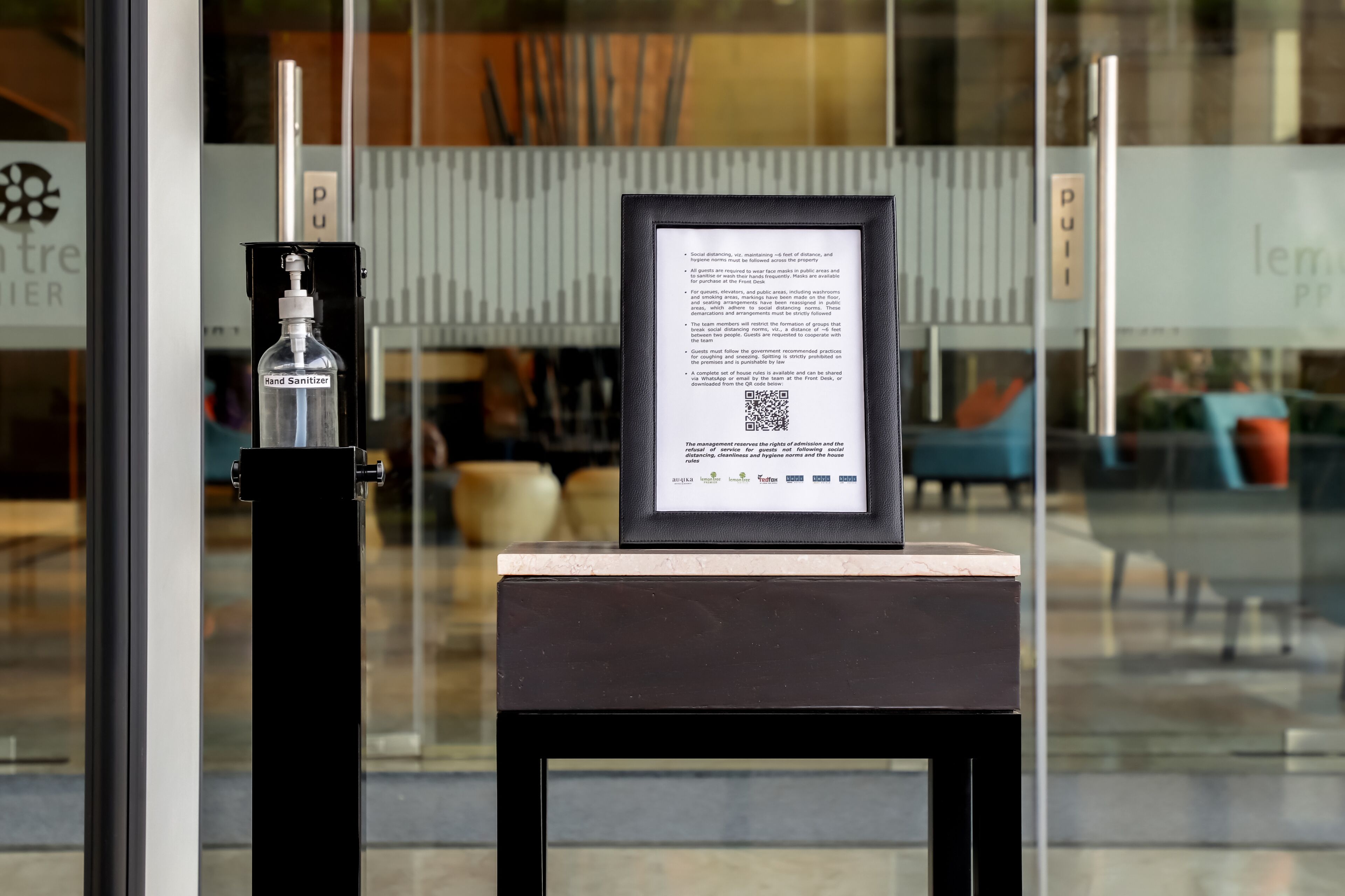 A hand sanitizer dispenser and a framed informational sign on a table outside a building with glass doors.