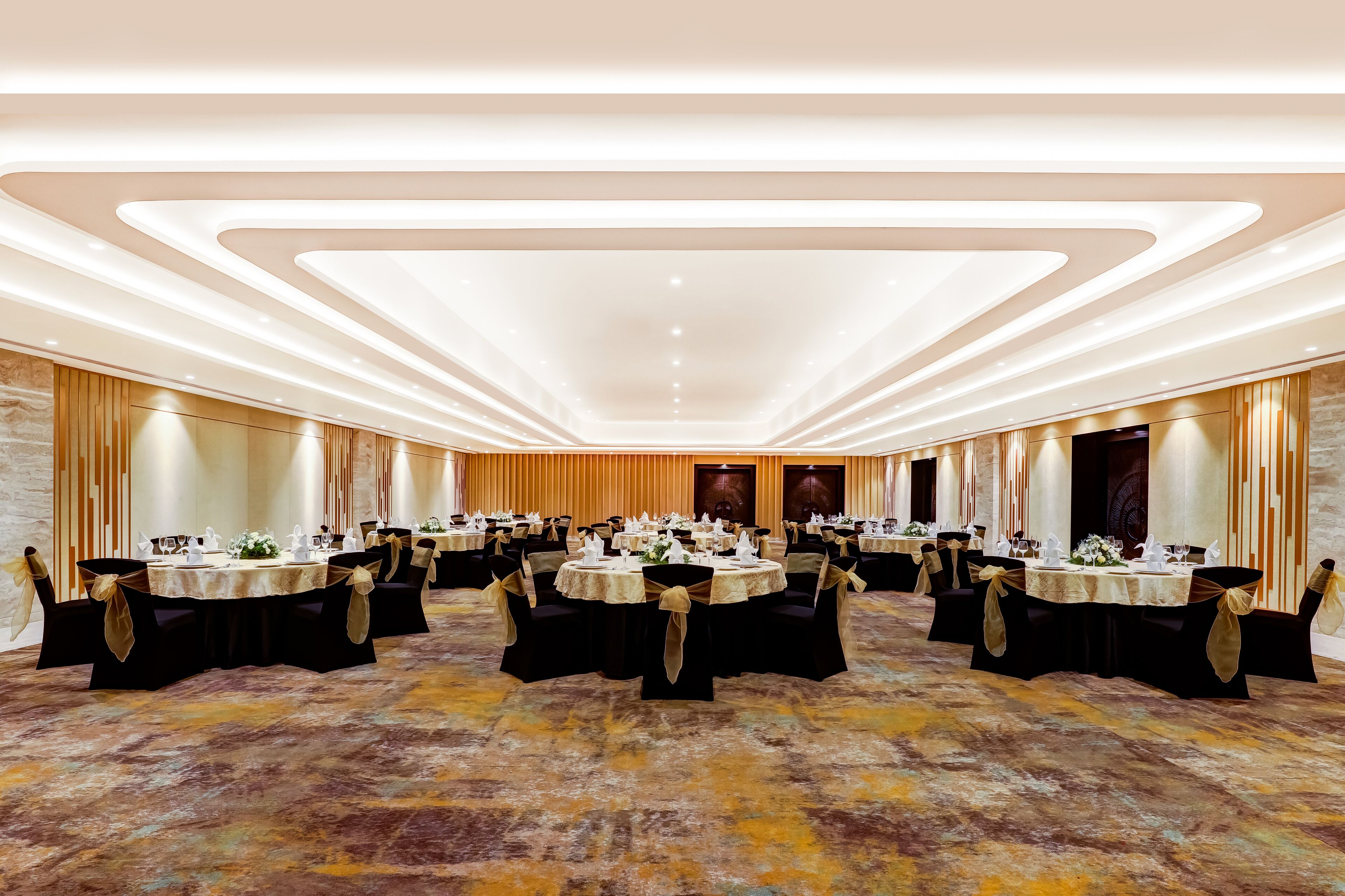 A spacious banquet hall with multiple round tables, each set with white tablecloths and black chairs, featuring gold sashes.