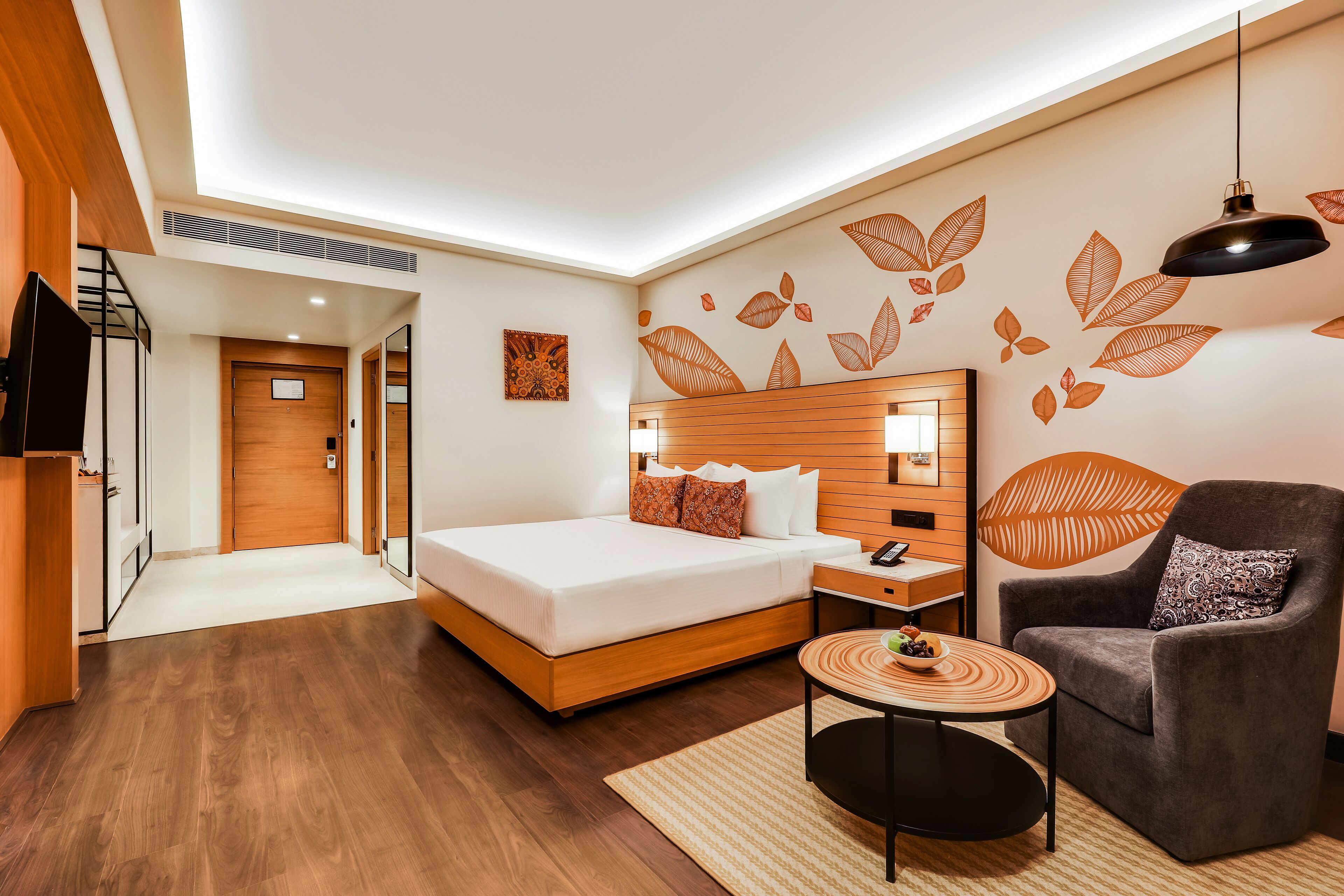A modern hotel room with a wooden bed, a grey armchair, a small round table, and a wall with leaf patterns.