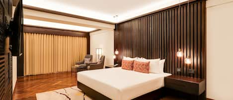 A modern hotel room with a large bed, wooden flooring, a television, a desk, and a wall-mounted light fixture.