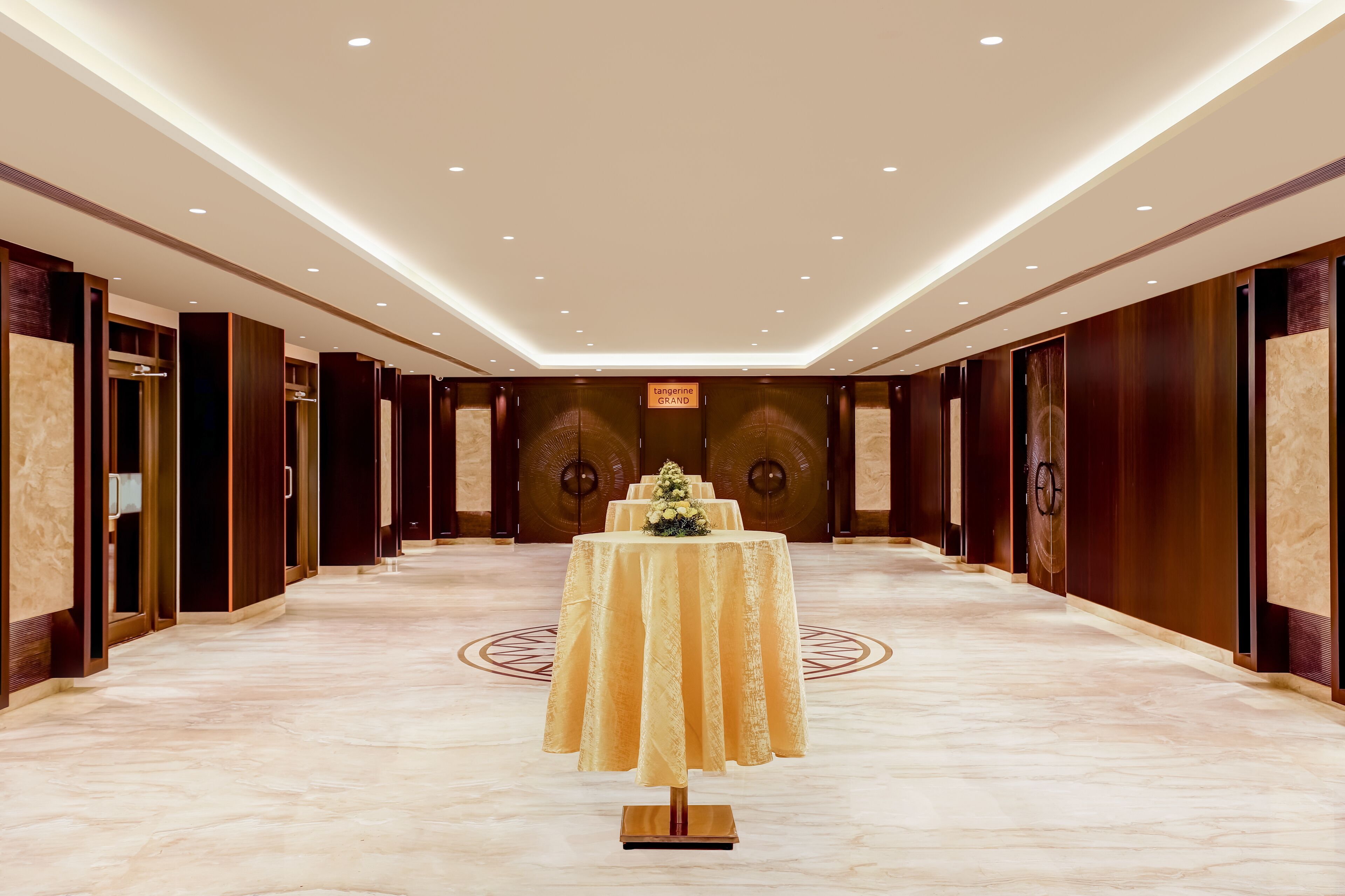 Un amplio pasillo de hotel con una mesa central decorada con flores, flanqueado por puertas de madera y un piso de mármol.