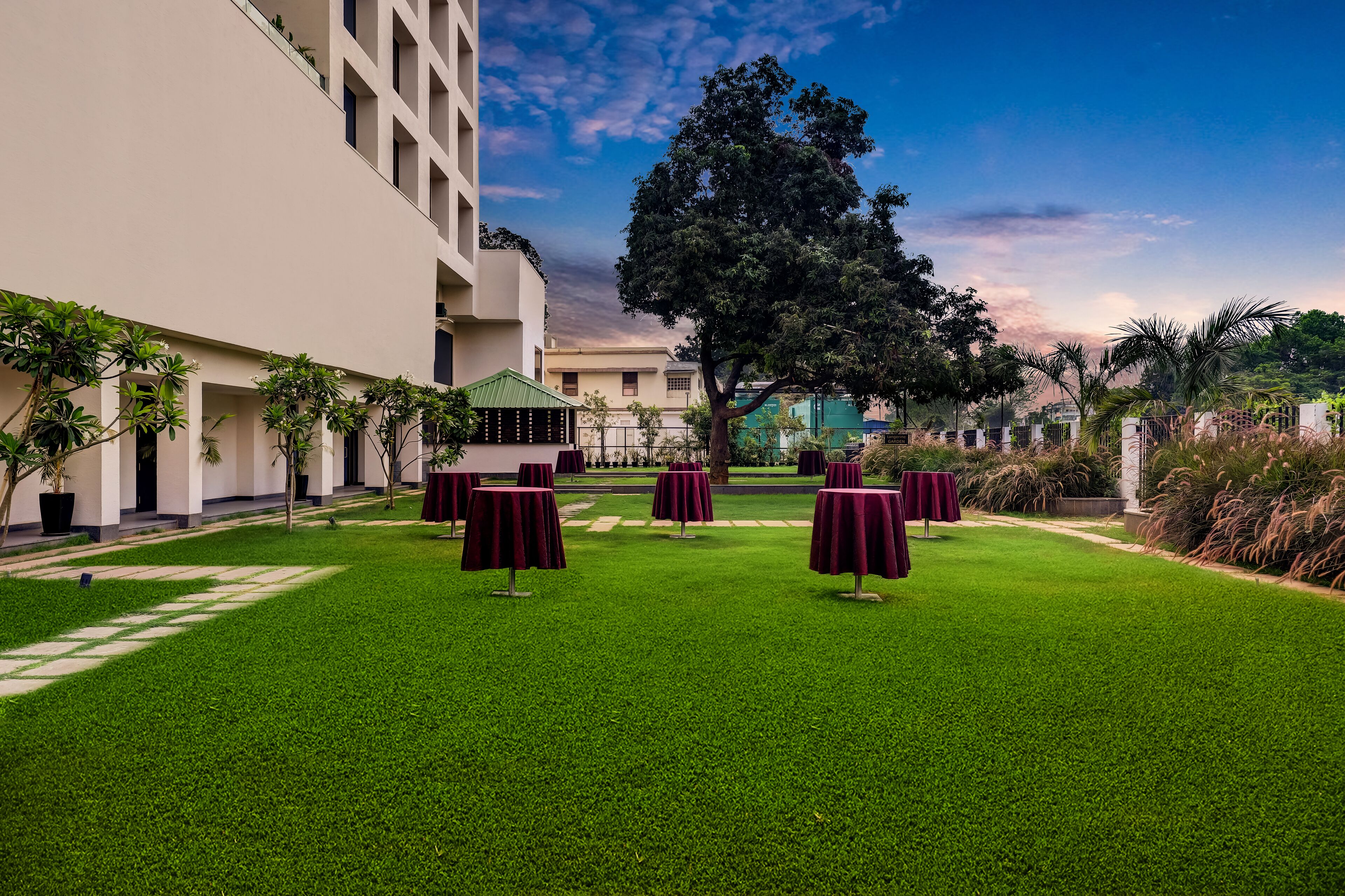 A courtyard with green grass, tables with red tablecloths, and a large tree.