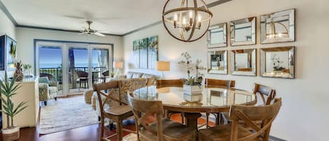 A dining area with a round table, wooden chairs, and a chandelier. There are multiple framed mirrors on the wall and a large window with a view.