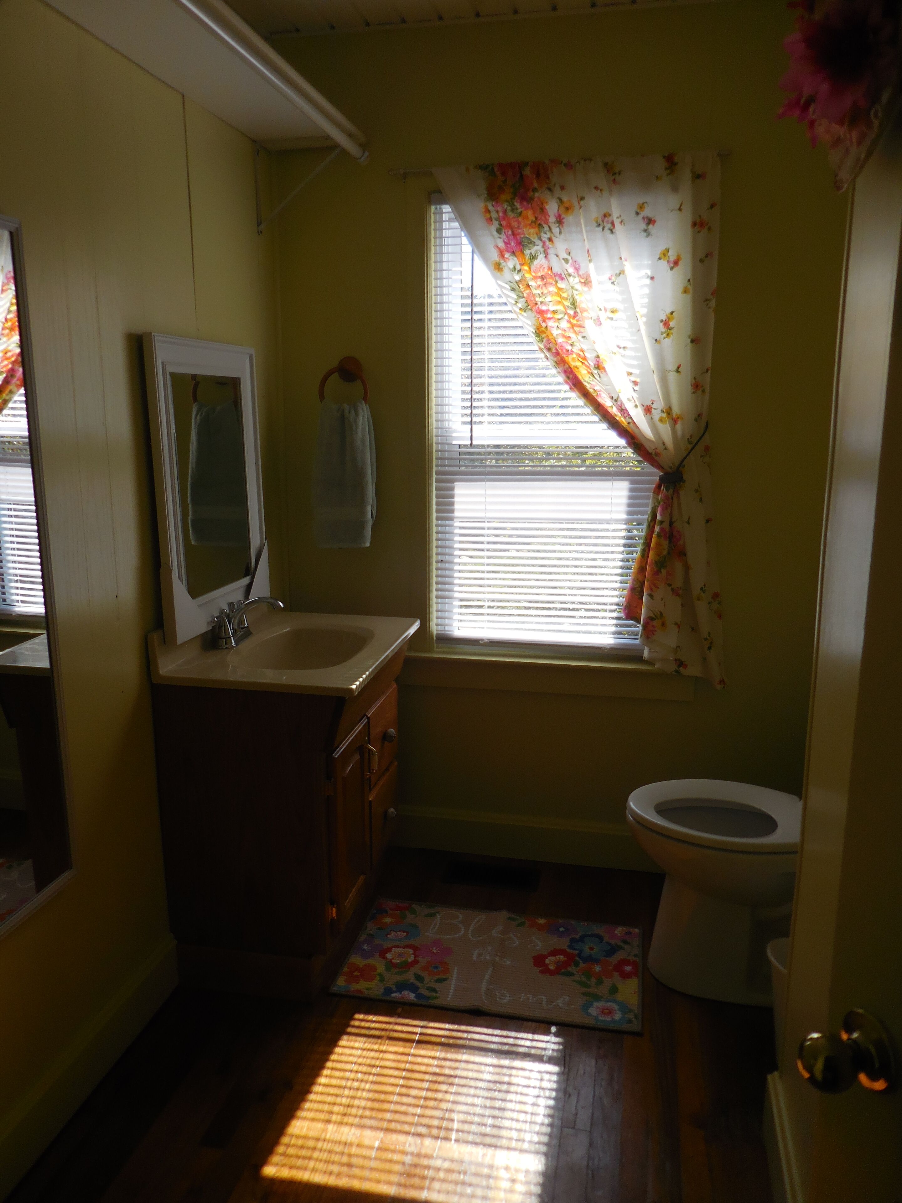 Een kleine badkamer met een wastafel, spiegel, toilet en raam met bloemen gordijnen.