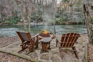A riverside campsite with a fire pit, wooden chairs, and a table.