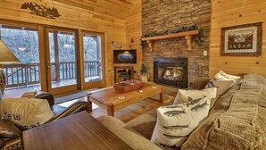 A cozy cabin interior with wooden walls, a stone fireplace, a television, and a balcony with a view of trees.