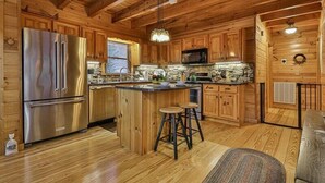 A wooden cabin interior with a kitchen featuring a stainless steel refrigerator, wooden cabinets, a stone countertop, and a bar stool.