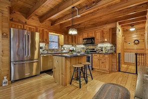 A wooden cabin interior with a kitchen featuring a stainless steel refrigerator, wooden cabinets, a stone countertop, and a bar stool.