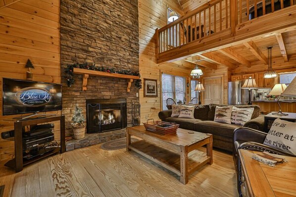 A cabin interior with a stone fireplace, wooden furniture, a television, and a balcony.