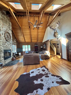 A log cabin interior with a stone fireplace, wooden ceiling beams, a ceiling fan, a mounted deer head, and a large cow hide rug.