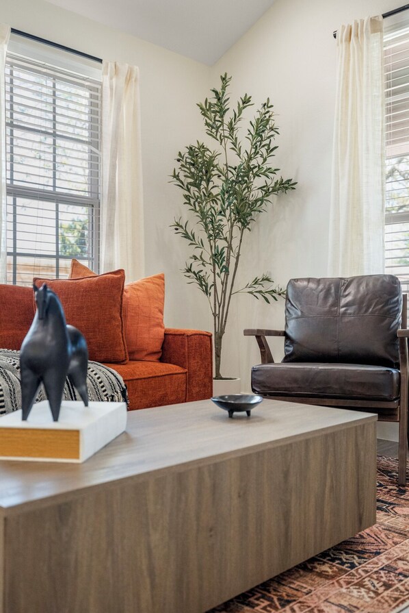A living room with a sofa, a coffee table, a chair, a potted plant, and a window with blinds.