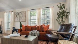 A living room with a sofa, coffee table, and a chair. There are windows with curtains, a cactus plant, and artwork on the wall. - Casa del rancho in the EE - 3 km from FIFA Fest (Houston)