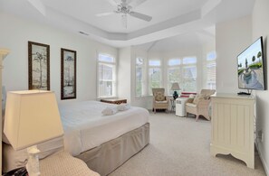 A bedroom with a bed, a chair, a desk, a TV, and large windows.