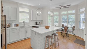 Una cocina moderna con armarios blancos, una isla central, taburetes de bar, un ventilador de techo y ventanas amplias.
