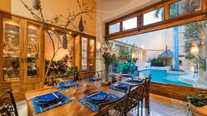 Uma sala de jantar com uma mesa de madeira posta para seis pessoas, rodeada de cadeiras, com vista para uma piscina e plantas tropicais.