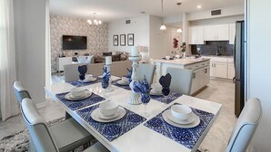 A modern dining area with a long table set for six, a kitchen in the background, and a living area with a sofa and TV. - Modern Condo 5mi to Disney, Foosball, Giant Water Slide, Free Water Park (Kissimmee)