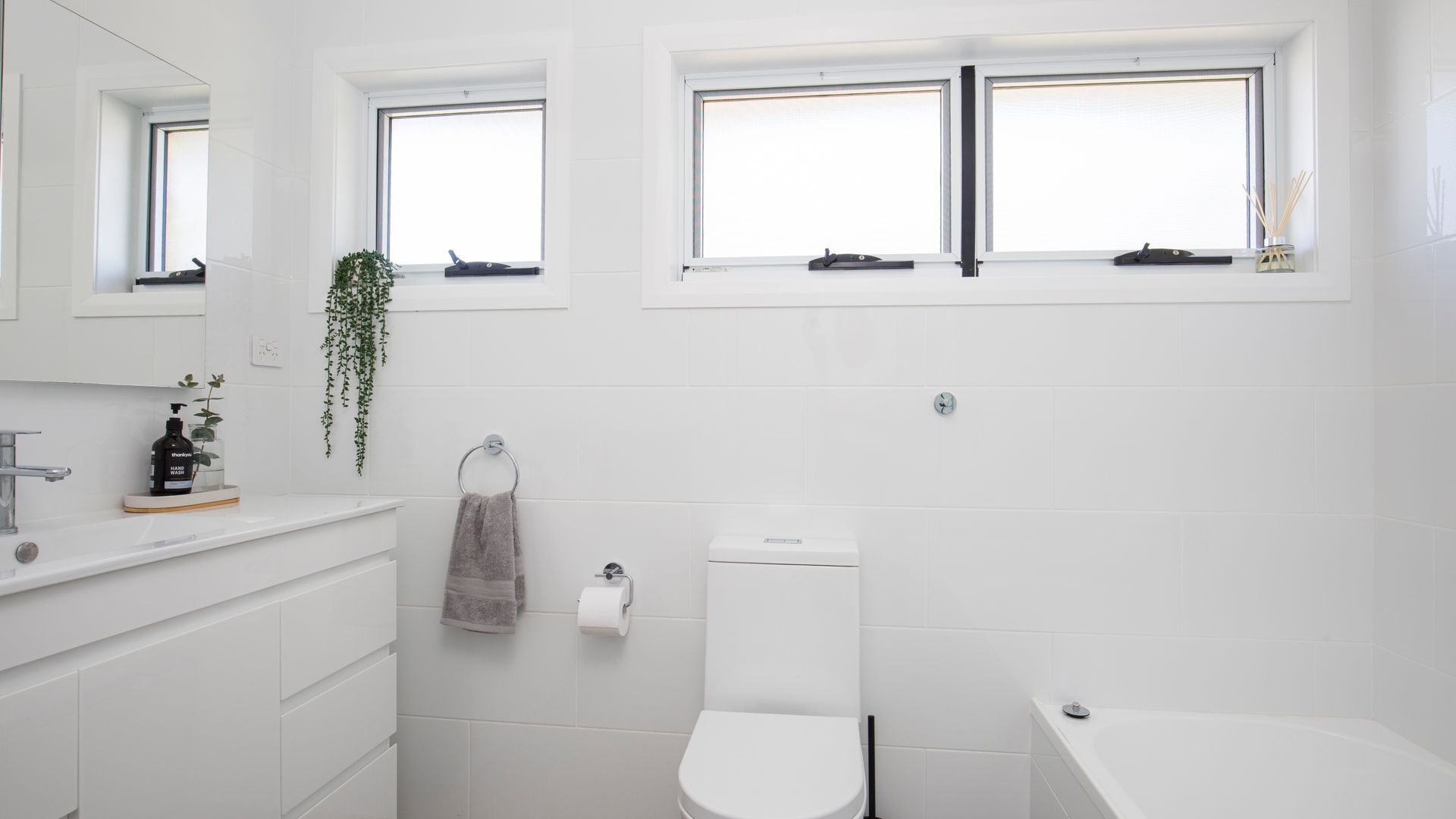 A modern bathroom with a toilet, sink, and bathtub. There are two windows providing natural light, and a plant is visible on the windowsill.