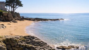 Uma paisagem costeira com uma praia arenosa, afloramentos rochosos e um edifício com árvores.