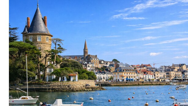 Chateau de Pornic, um edifício histórico de pedra com uma torre, localizado próximo a um porto com barcos e uma igreja ao fundo.