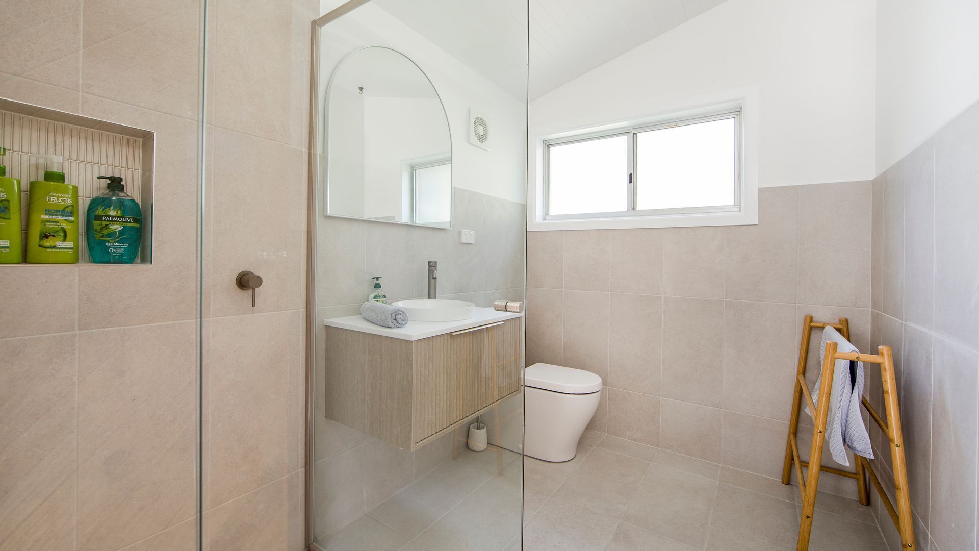 A modern bathroom with a glass shower enclosure, a wooden vanity, a white toilet, and a wooden ladder with towels.