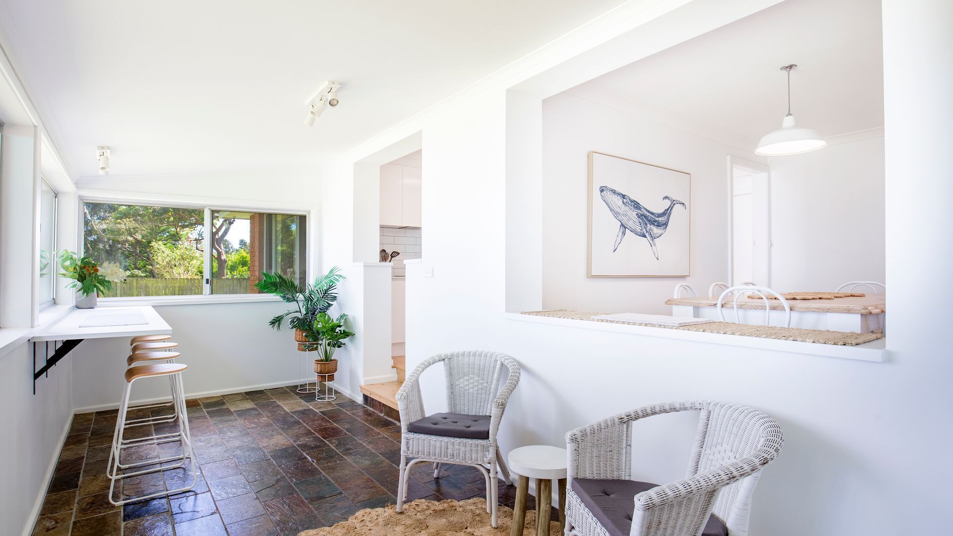 Una cocina luminosa con armarios blancos, una zona de comedor con sillas de mimbre y un gran ventanal con vistas a zonas verdes.