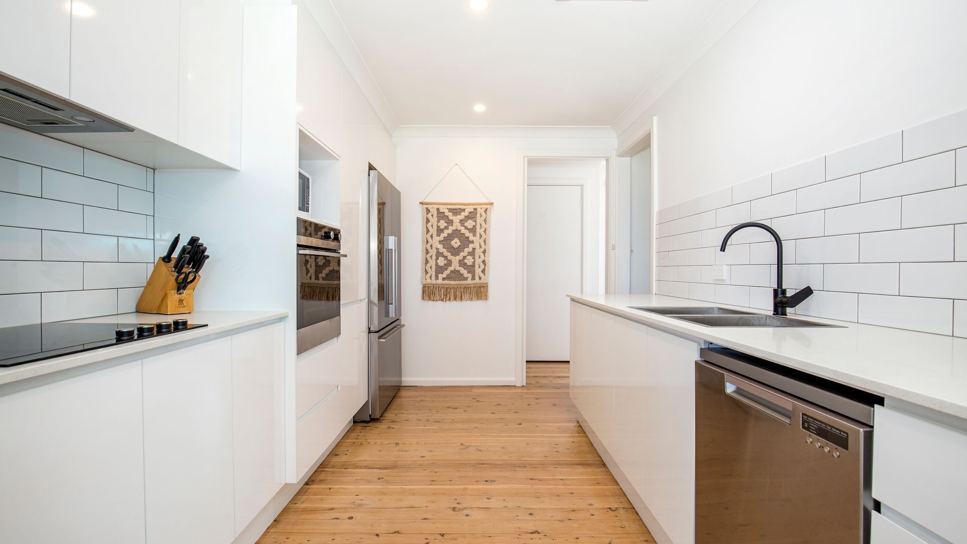 A narrow kitchen with white cabinets, a wooden floor, a stainless steel dishwasher, and a hanging decorative piece.